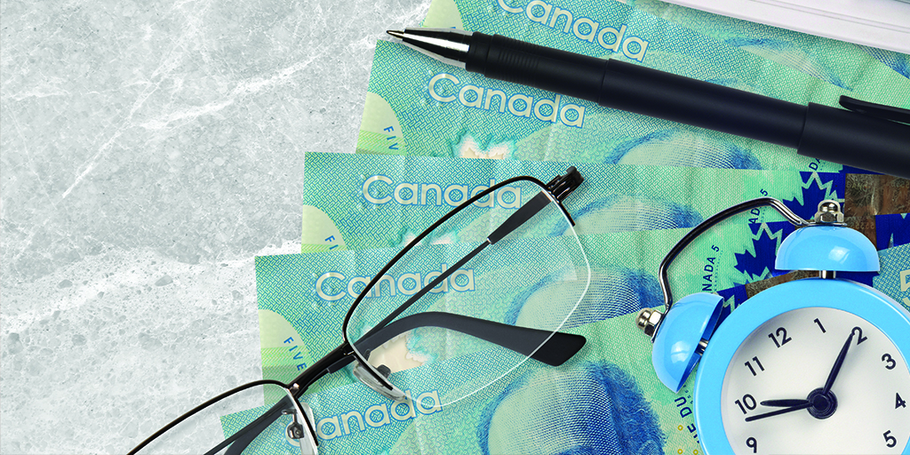 An image of glasses, a pen, and an alarm clock on top of five dollar Canadian bills