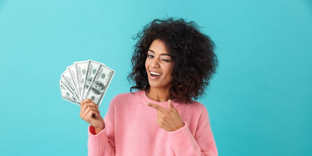 An image of a black woman in a pink shirt holding and pointing at fanned out American one hundred dollar bills in front of a blue background.